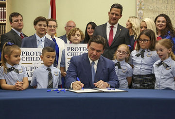 Photo montrant le gouverneur de Floride, Ron DeSantis signant le projet de loi Don't Say Gay. Derrière lui, un groupe d'enfants et d'adultes portant des pancartes "Protect Childrens"