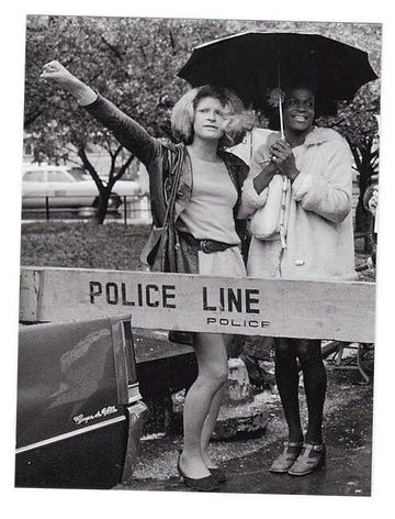 Photo en noir et blanc montrant Sylvia Rivera et Marsha P.Johnson sous un parapluie, derrière une barrière de police. Sylvia brandit son point tandis que Marsha serre le parapluie, en souriant. 