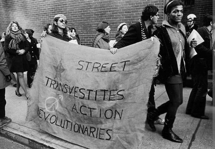 Photo d'archives en noir et blanc représentant Marsha tenir une banderole "STREET TRANSVESTITES ACTION REVOLUTIONARIES"